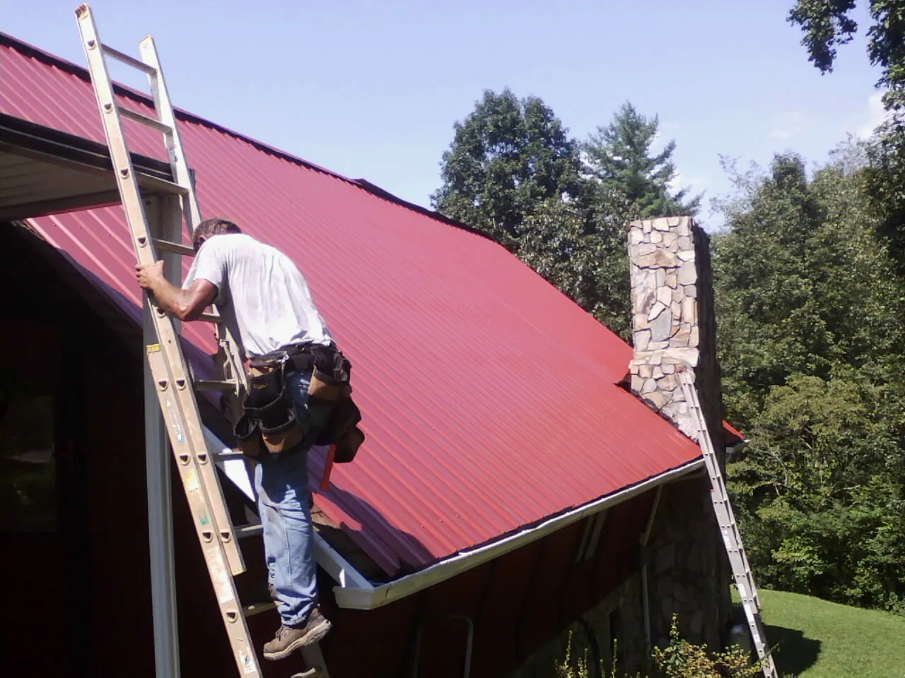 Roofer on a ladder beside a red metal roof and stone chimney — Soffit & Fascia Repair by Roofing Brothers in Macon